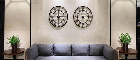 A modern living room with a grey sofa, two wall-mounted clocks, and two side tables with potted plants.