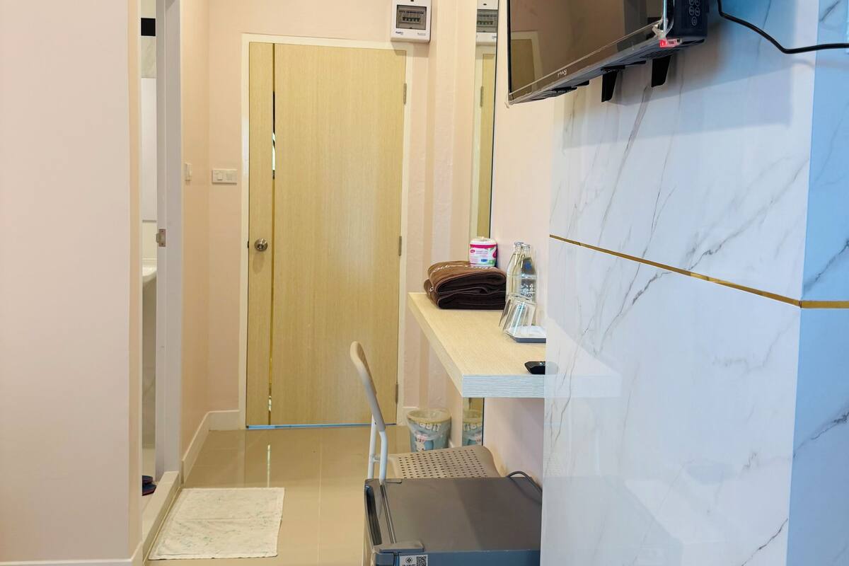 A compact hotel room with a wooden door, a small desk with a chair, a flat-screen TV mounted on the wall, and a refrigerator.