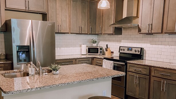 A kitchen with wooden cabinets, a stainless steel refrigerator, a microwave, a stove, and two stools.