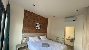Un dormitorio moderno con pared de ladrillo decorativa, una cama grande con ropa de cama blanca, una mesita de noche con lámpara, y un baño con lavamanos y espejo.