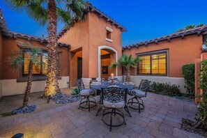 A patio with a round table and chairs, palm trees, and a building with a tiled roof.