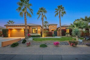 A residential property with a two-story house, a garage, and a well-maintained garden with palm trees and flowering plants.