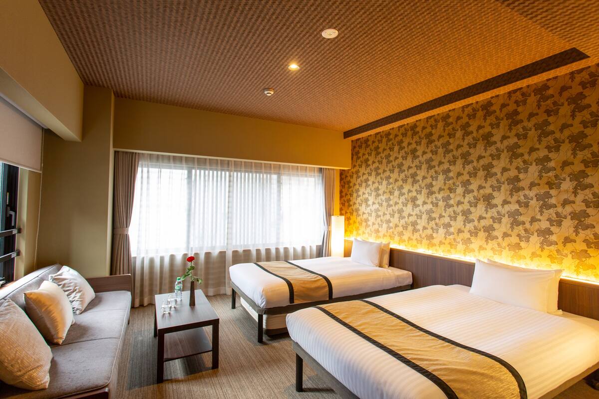 A hotel room with two beds, a sofa, a small table, and a large window with curtains.
