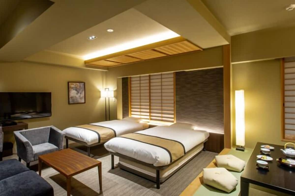 A hotel room with two beds, a TV, a sofa, a table, and a window with blinds.