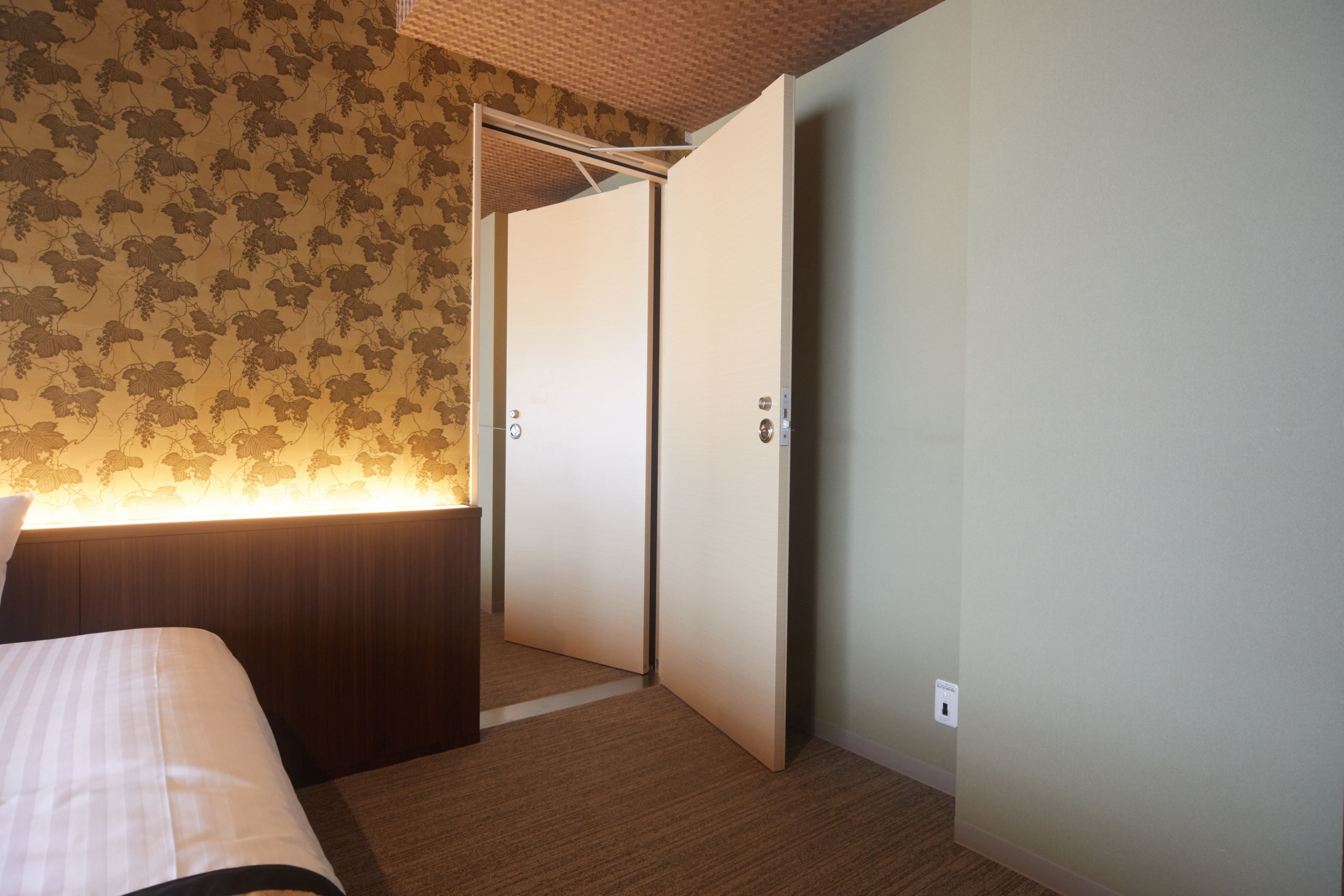 A hotel room with a bed, a nightstand, a lamp, a wardrobe, and a carpeted floor.
