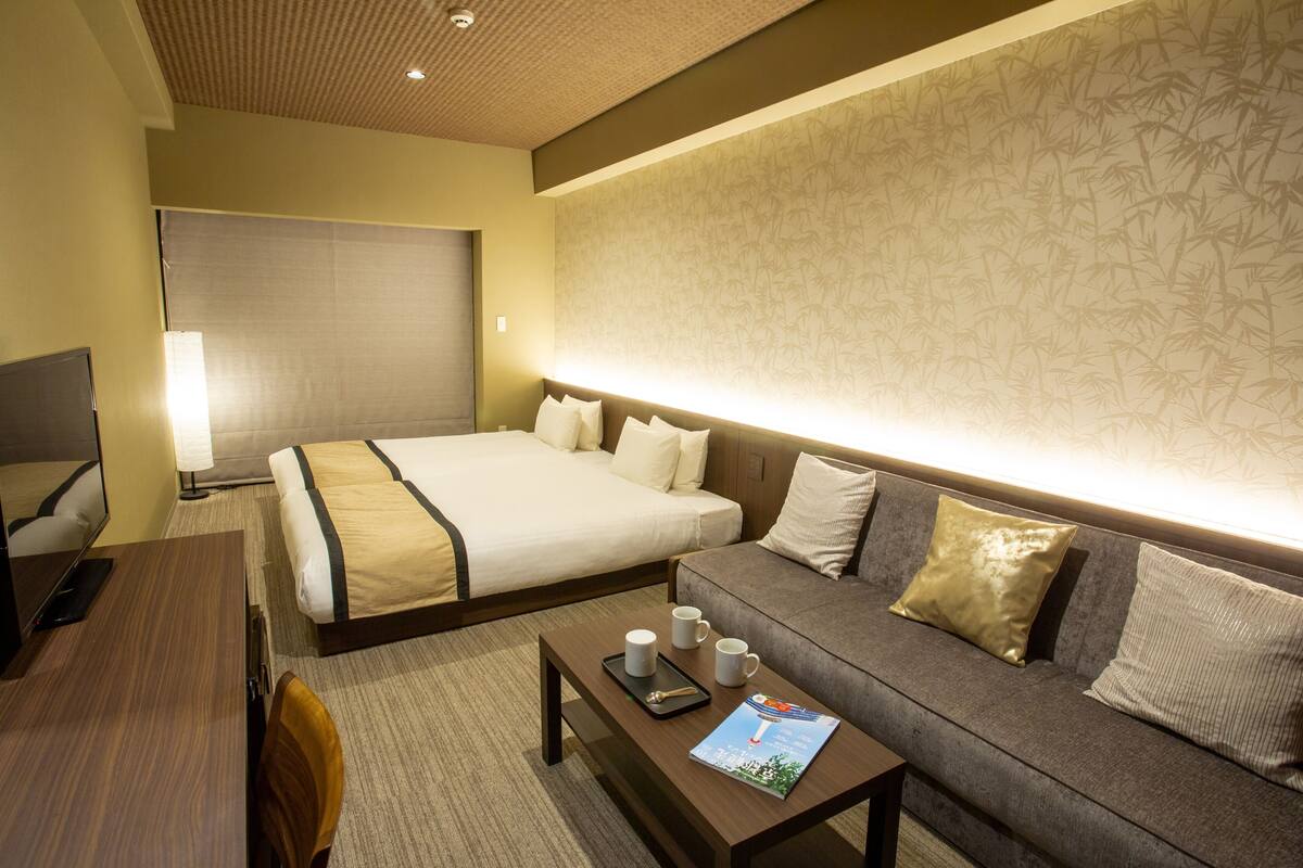 A hotel room with a bed, a sofa, a coffee table with a magazine, and a TV on a stand.
