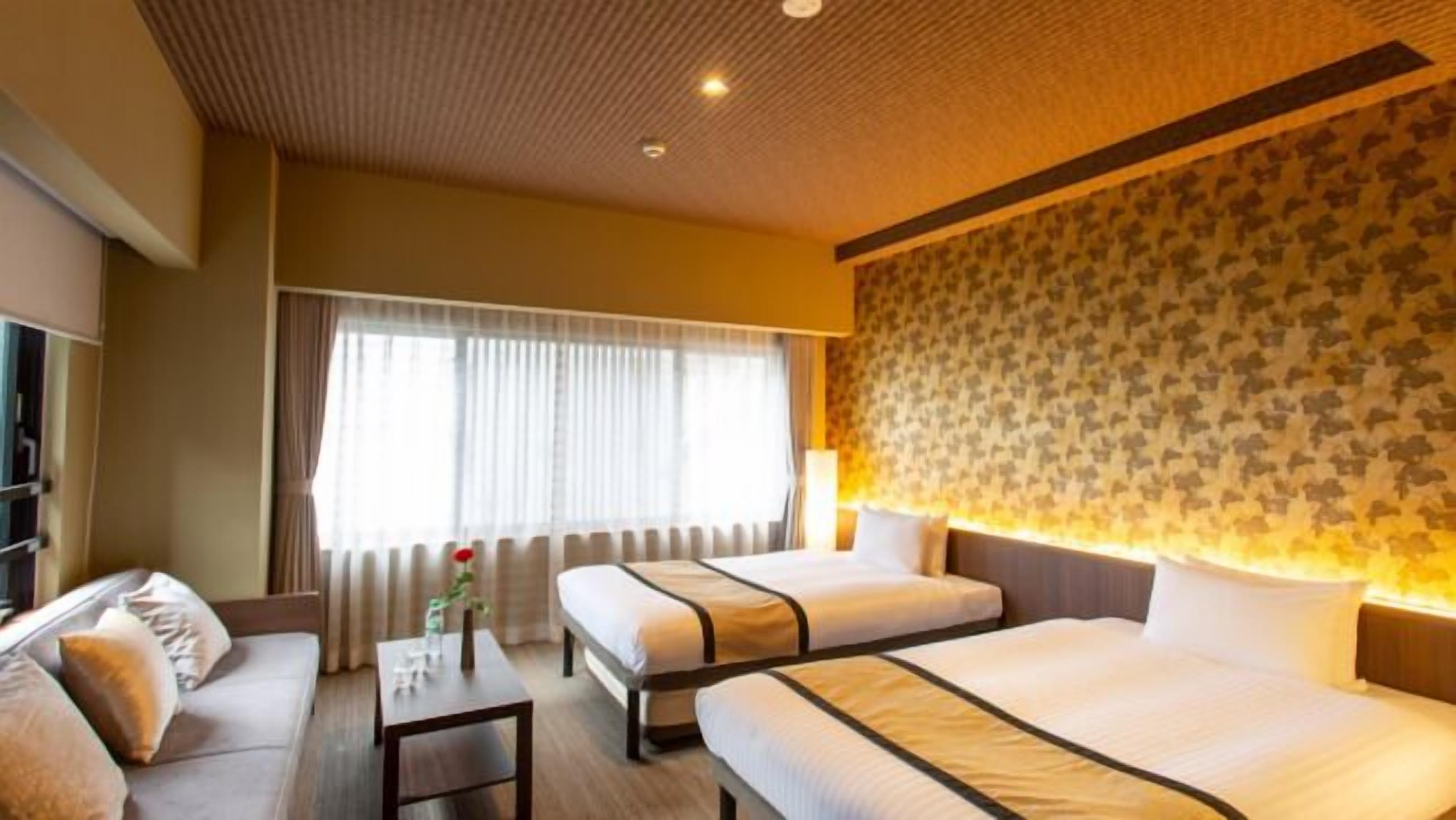 Une chambre d’hôtel avec deux lits, un canapé, une petite table, une lampe et une fenêtre avec des rideaux.