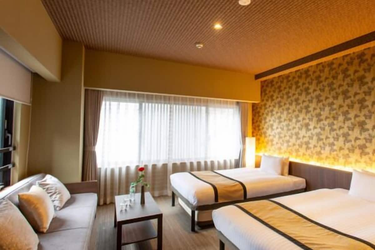 A hotel room with two beds, a sofa, a small table, a lamp, and a window with curtains.