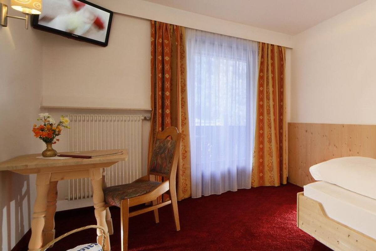 Quarto de hotel com cabeceira de madeira, cama com lençóis brancos, pequena mesa de trabalho com um vaso de flores, uma cadeira e uma televisão na parede.