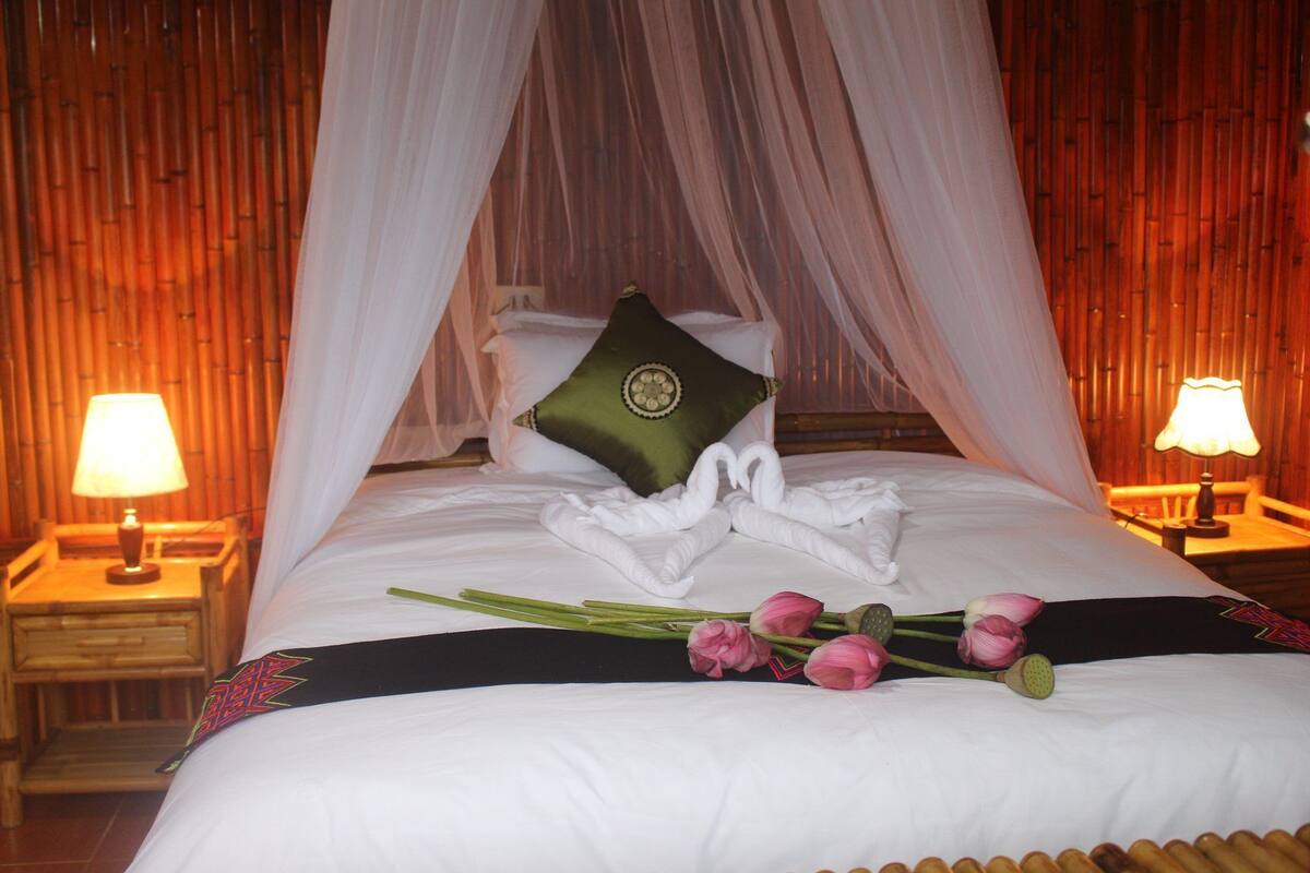 A bed with white linens, a green pillow, and a decorative swan towel arrangement. There are two bedside lamps and a bouquet of pink flowers on the bed.