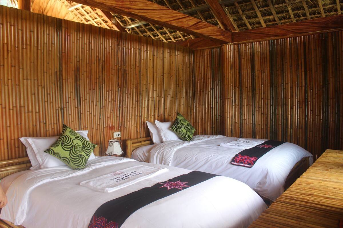 A room with two beds, each with white linens and decorative pillows. The walls are covered with vertical bamboo panels, and the ceiling has a wooden structure with a thatched roof.