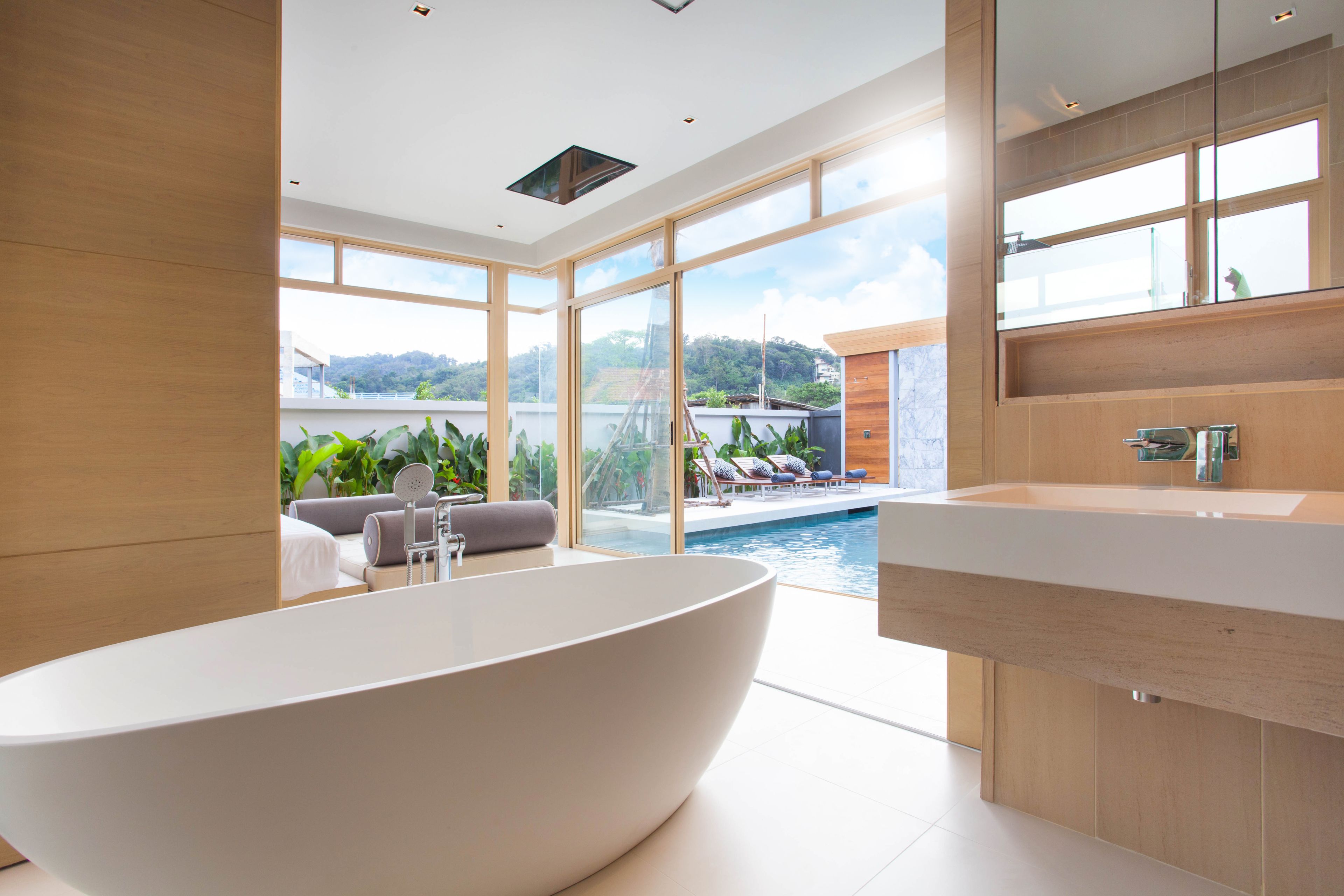 A modern bathroom with a freestanding bathtub, large windows, and a view of a pool and outdoor seating area.