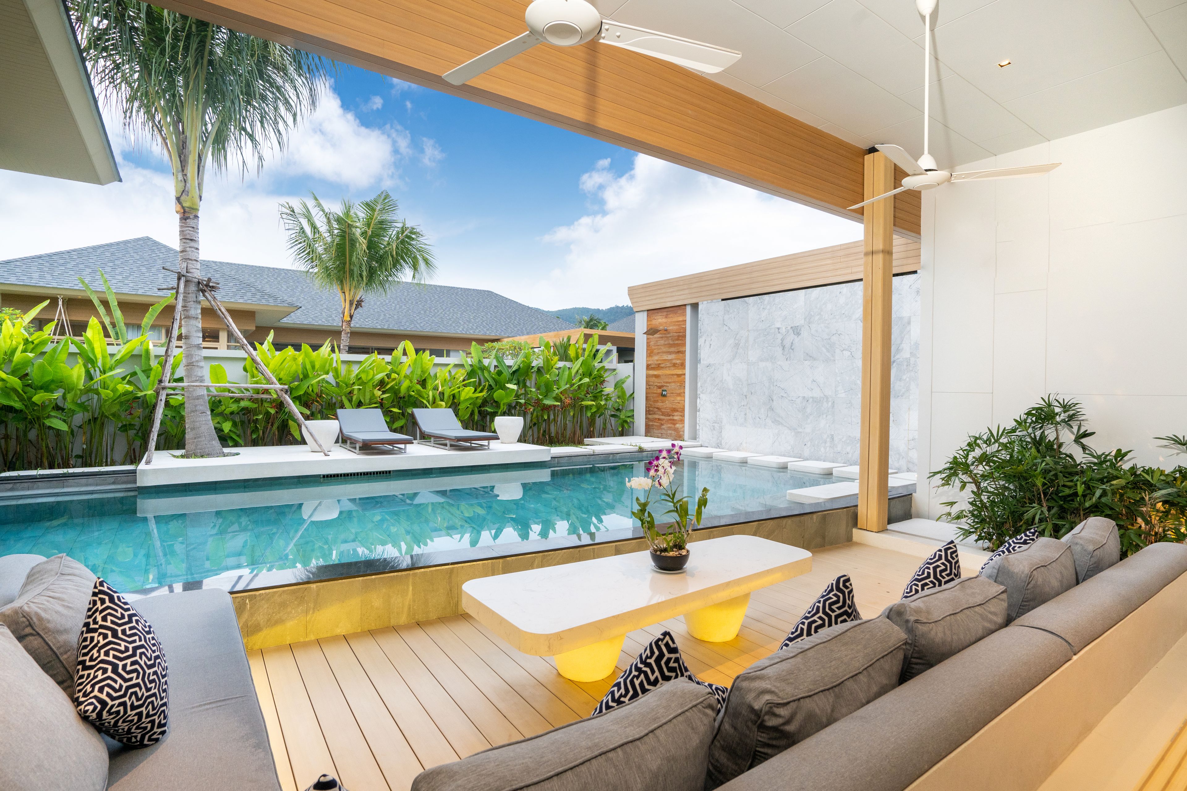 A modern outdoor pool area with a wooden deck, a sofa with patterned cushions, and a ceiling fan.