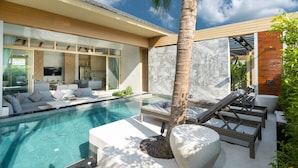 A modern outdoor pool area with a lounge chair, a palm tree, and a well-lit living space with a TV.