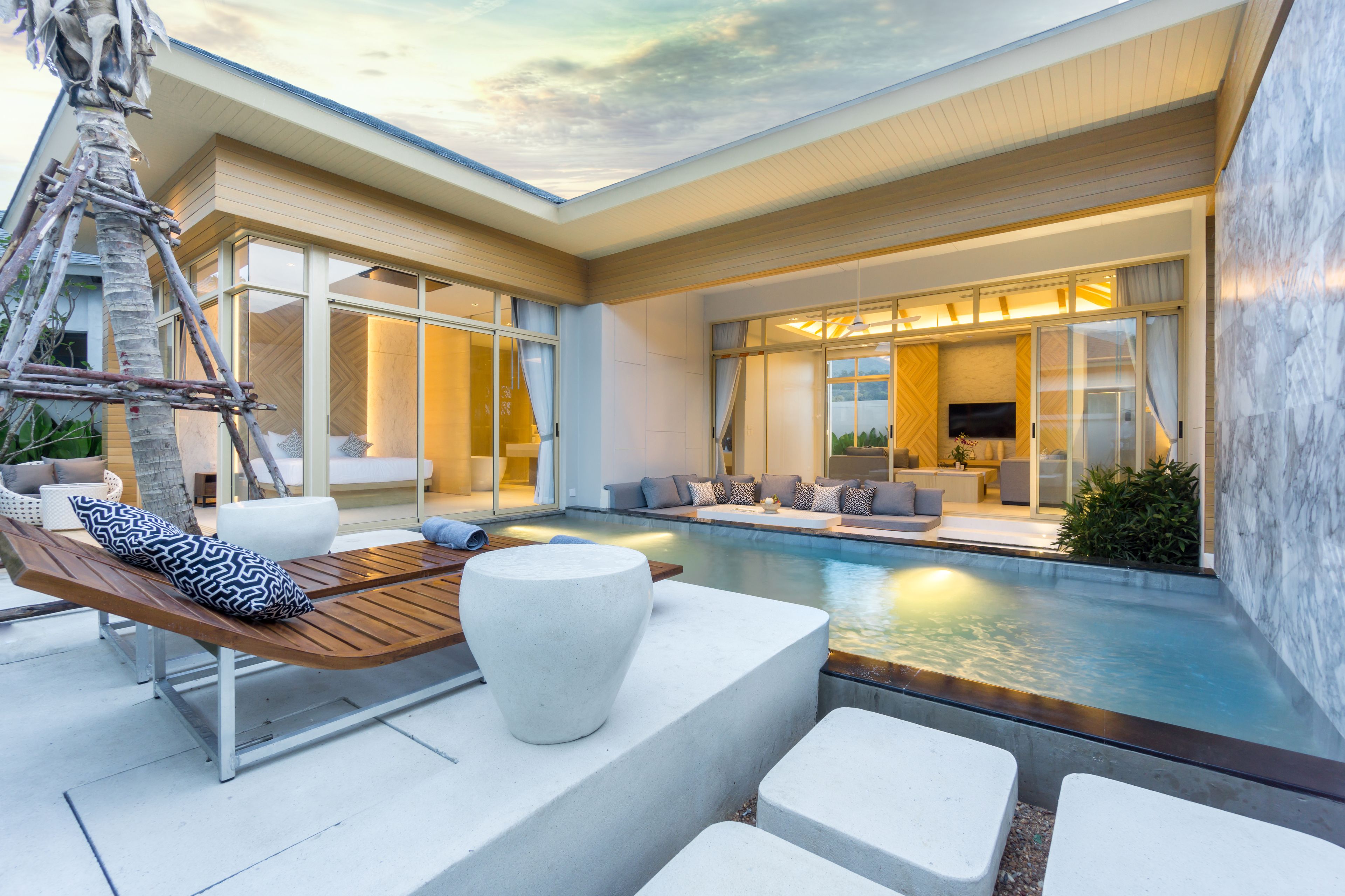 A modern outdoor pool area with a wooden lounge chair, white seating, and a stone planter.