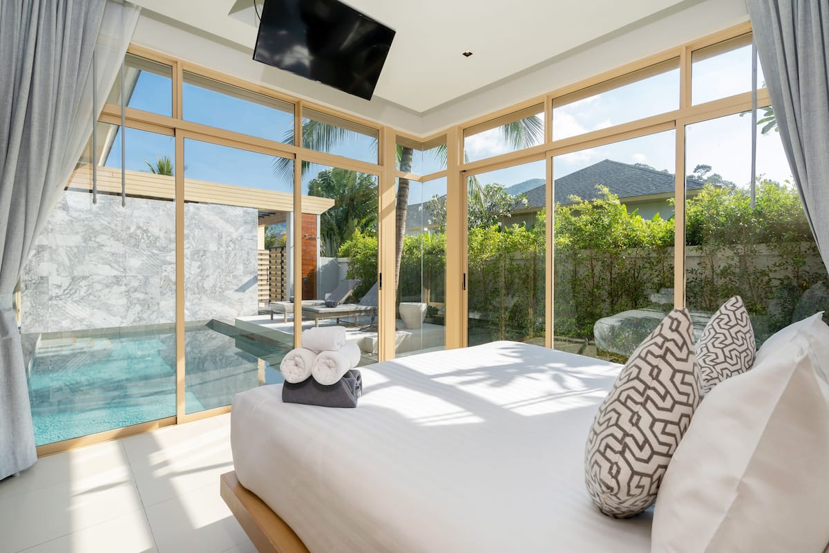 A modern bedroom with a large bed, a TV, and a view of a swimming pool and greenery.
