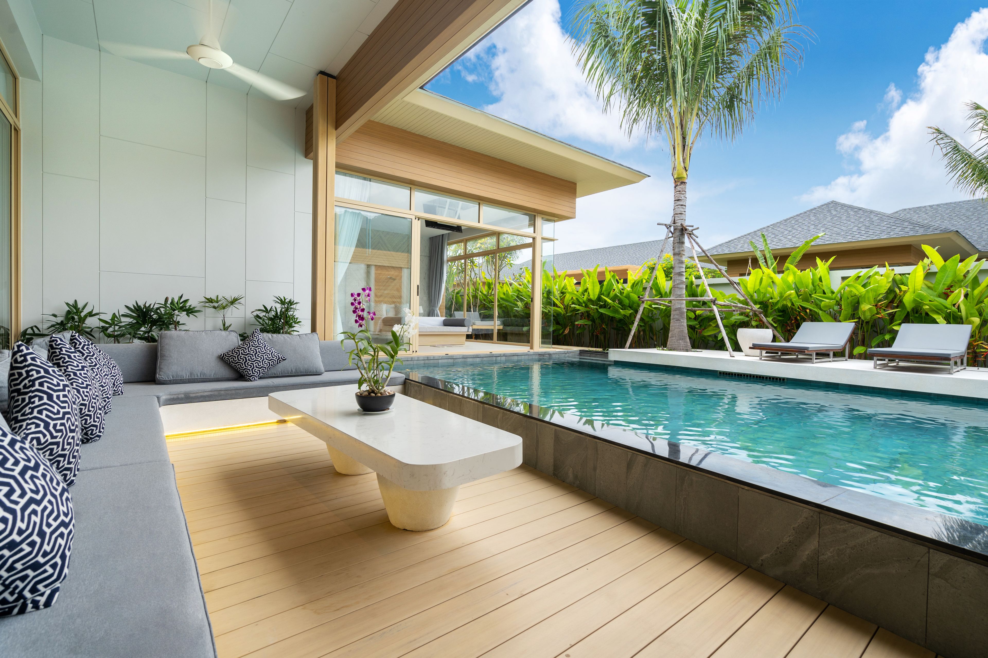 A modern outdoor pool area with a wooden deck, a seating area with a sofa and a coffee table, and lush greenery.