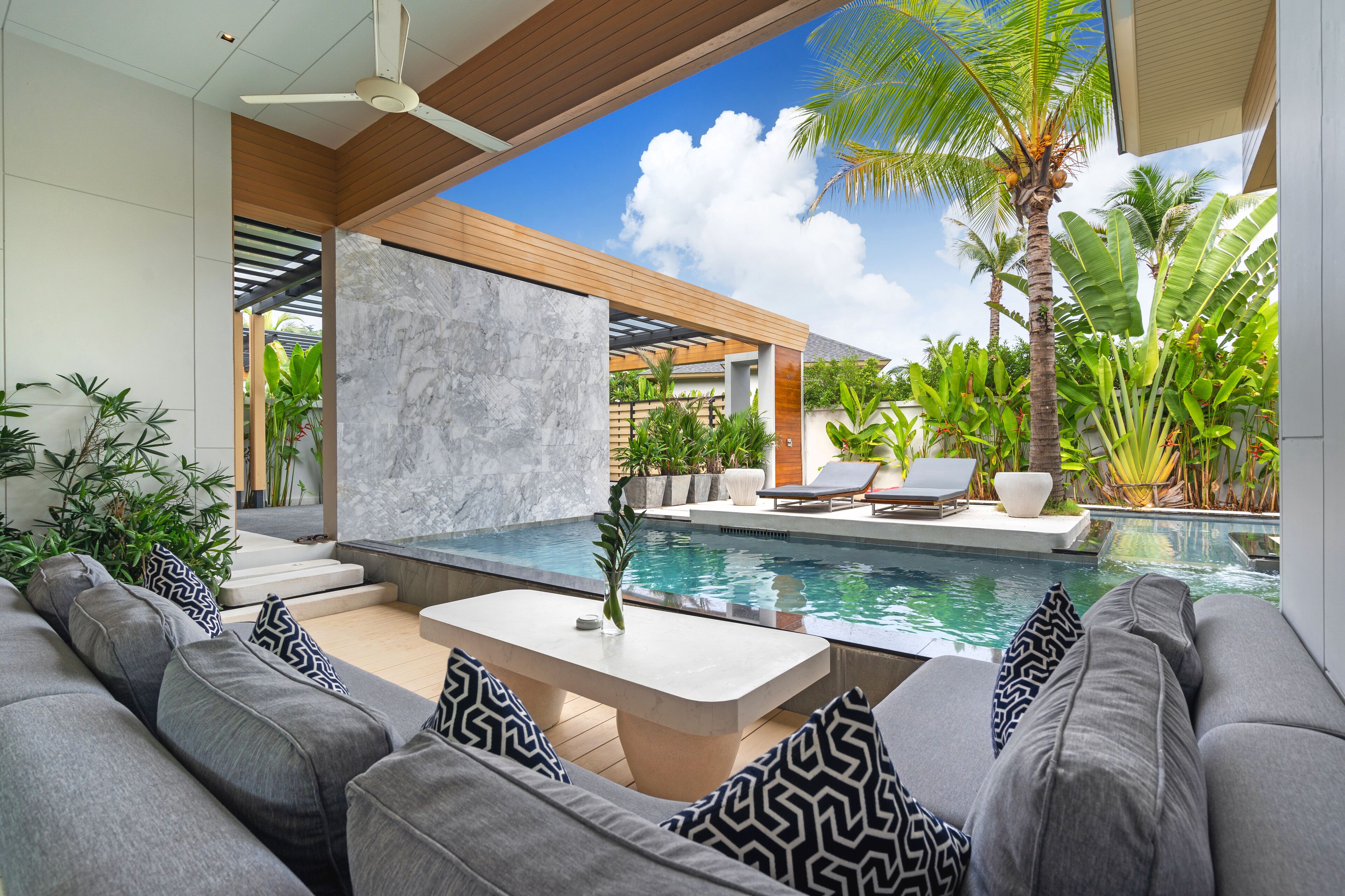 A modern outdoor living area with a pool, grey sofas, and tropical plants.