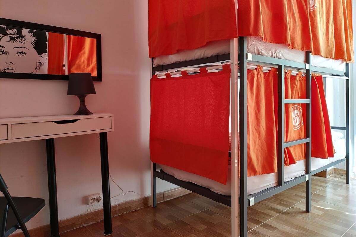A bunk bed with orange bedding, a desk with a lamp, and a wall-mounted artwork.