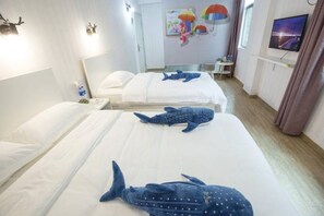 Quarto de hotel com duas camas, cada uma com um travesseiro em formato de baleia azul. Há uma televisão na parede e uma mesa pequena com uma luminária.