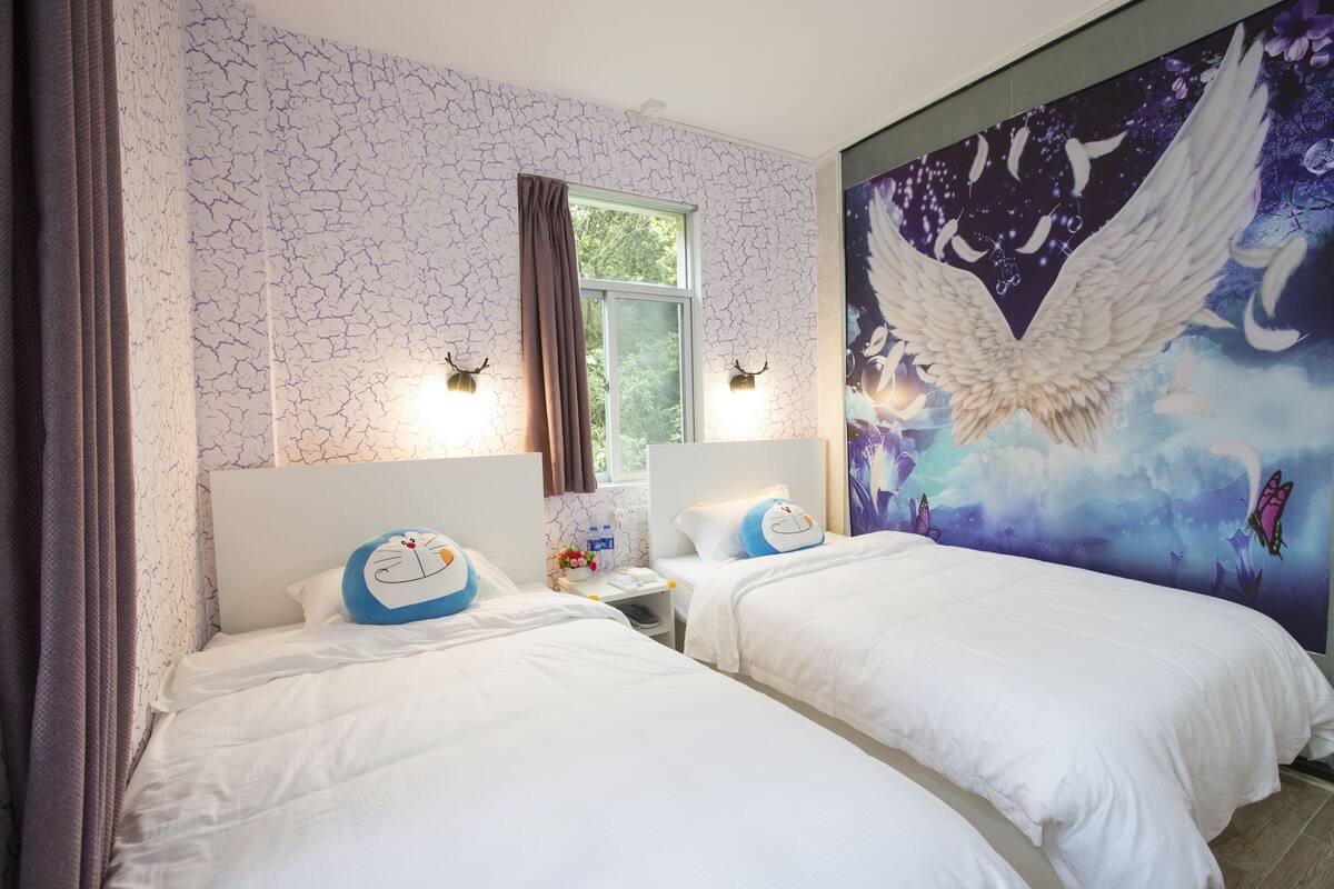 A hotel room with two beds, a window, and a mural of an angel on the wall.
