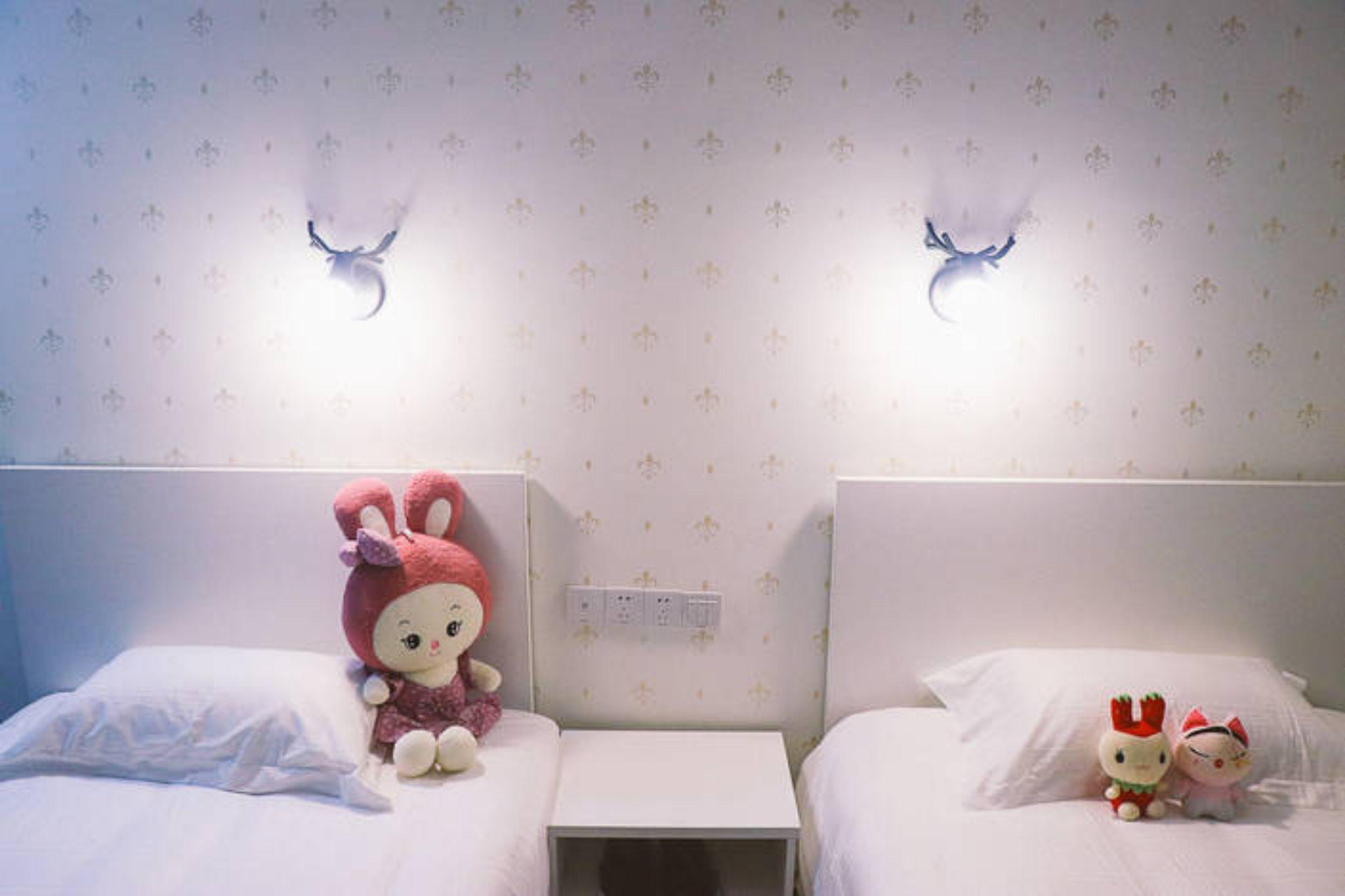 Two single beds with white bedding and floral wallpaper. Each bed has a small bedside table and a wall-mounted light fixture.