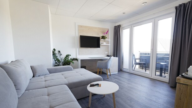 Ein modernes Wohnzimmer mit einer grauen Eckcouch, einem kleinen runden Tisch, einem flachbildfernseher an der Wand und einem Balkon mit Sitzgelegenheit.