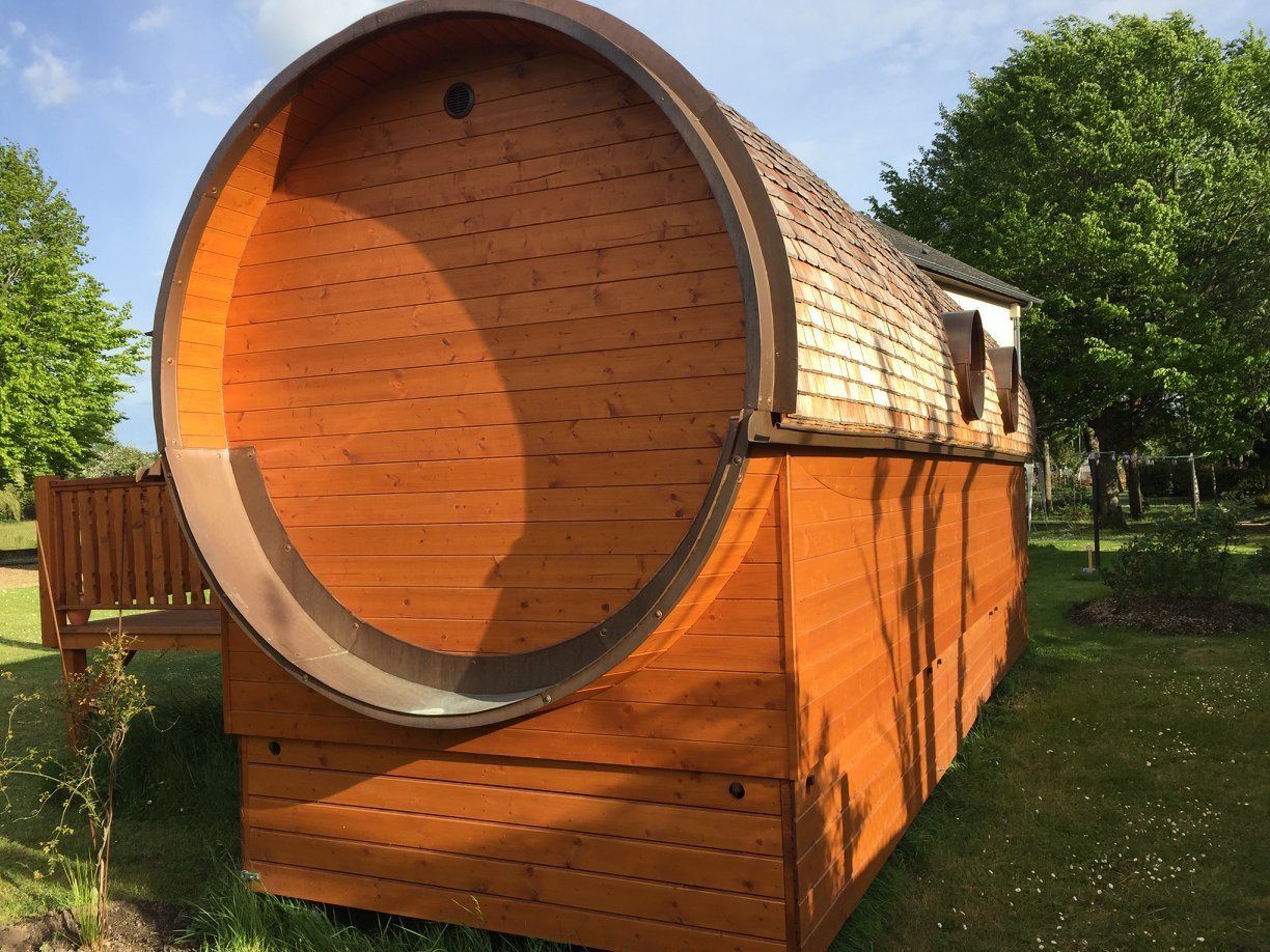 A wooden barrel-shaped building with a large circular opening, a bench, and a grassy area with trees.