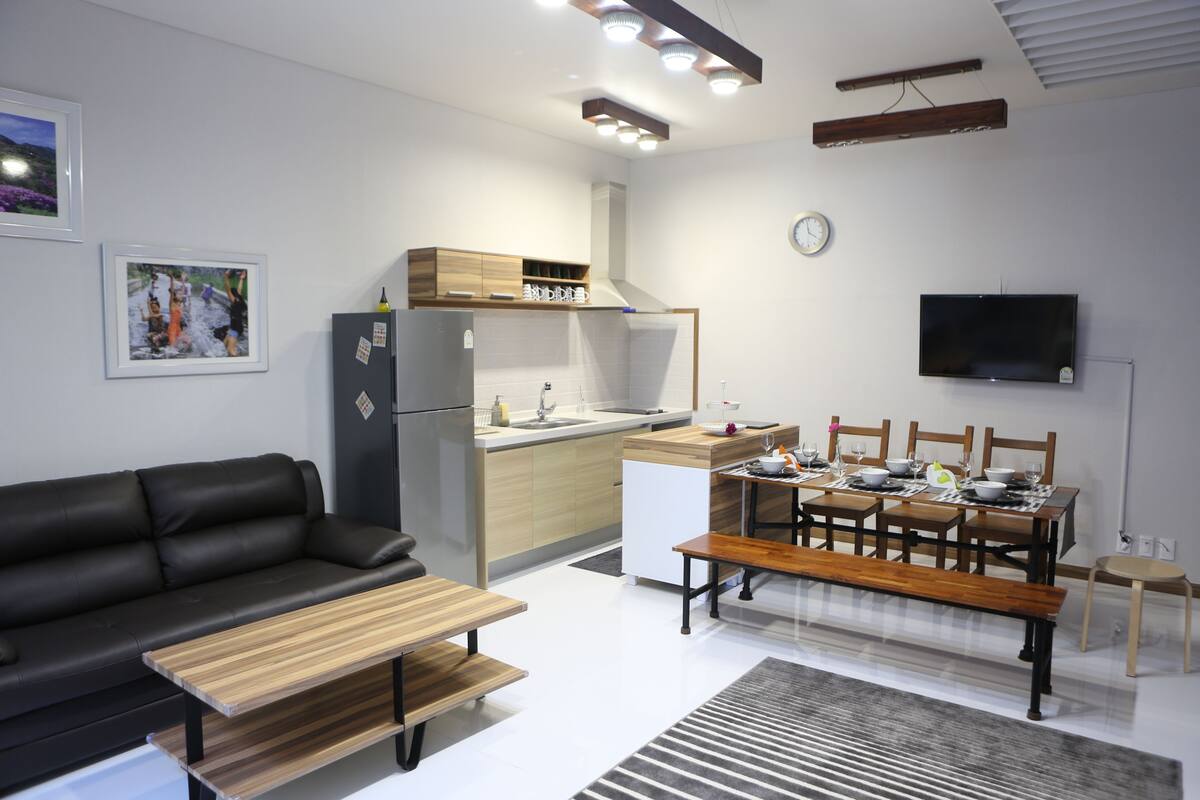 Moderni olohuone, jossa on sohva, ruokapöytä tuoleineen, keittiöalue ja seinälle asennettu televisio.