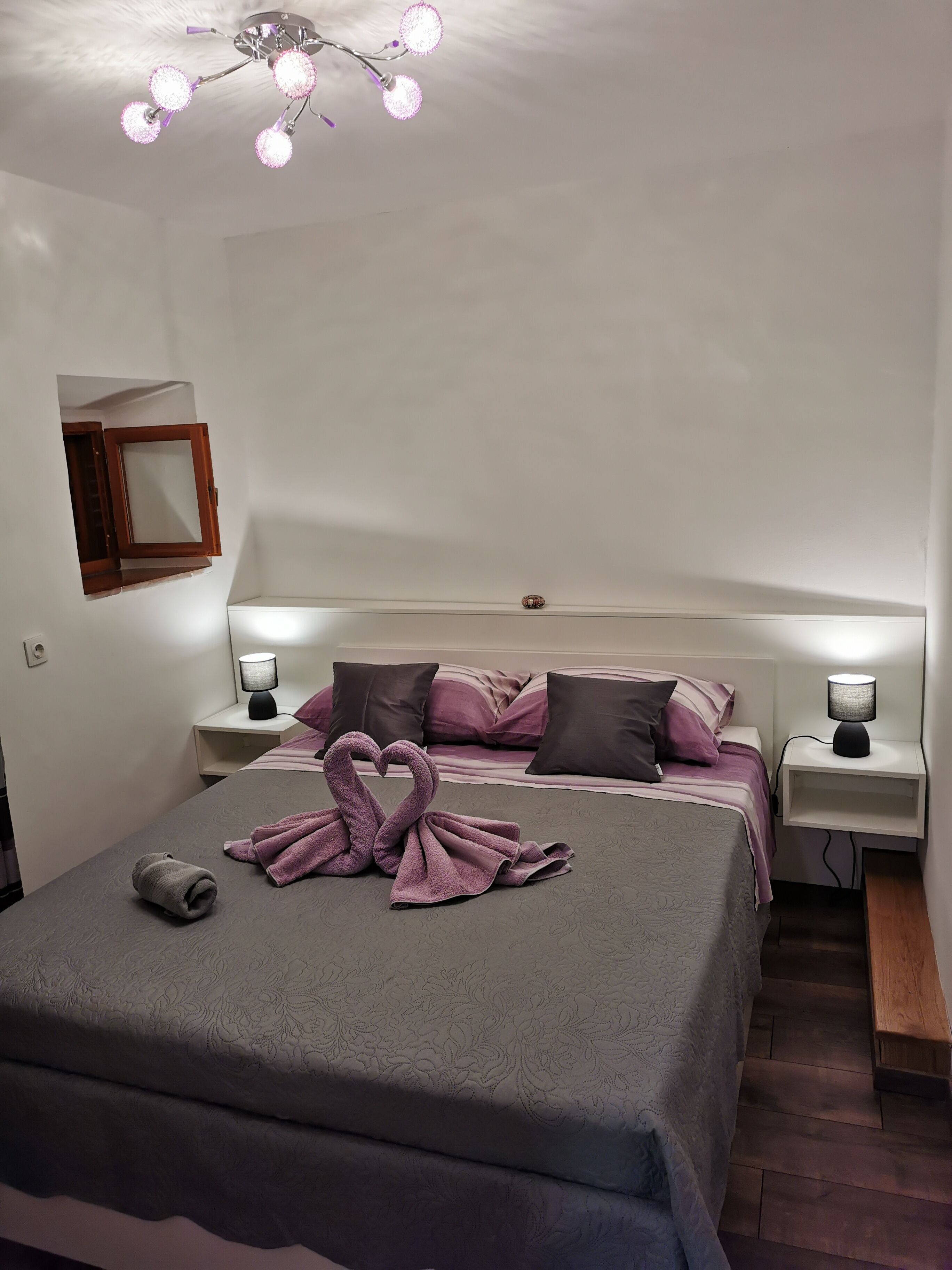Una cama bien hecha con una decoración de toallas en forma de cisne, lámparas de noche y un aplique en el techo.