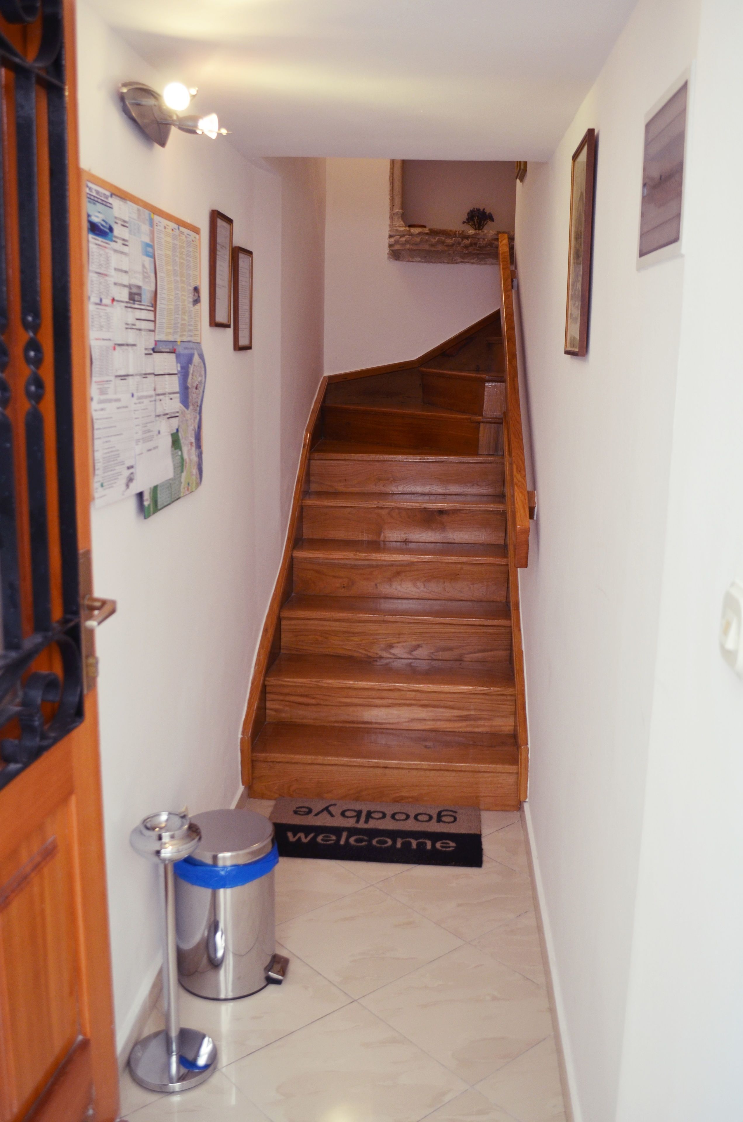 A narrow hallway with a wooden staircase leading to a landing. A metal trash bin and a welcome mat are visible on the floor.