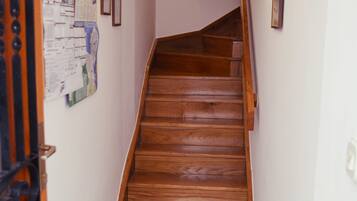 A narrow hallway with a wooden staircase leading to a landing. A metal trash bin and a welcome mat are visible on the floor.