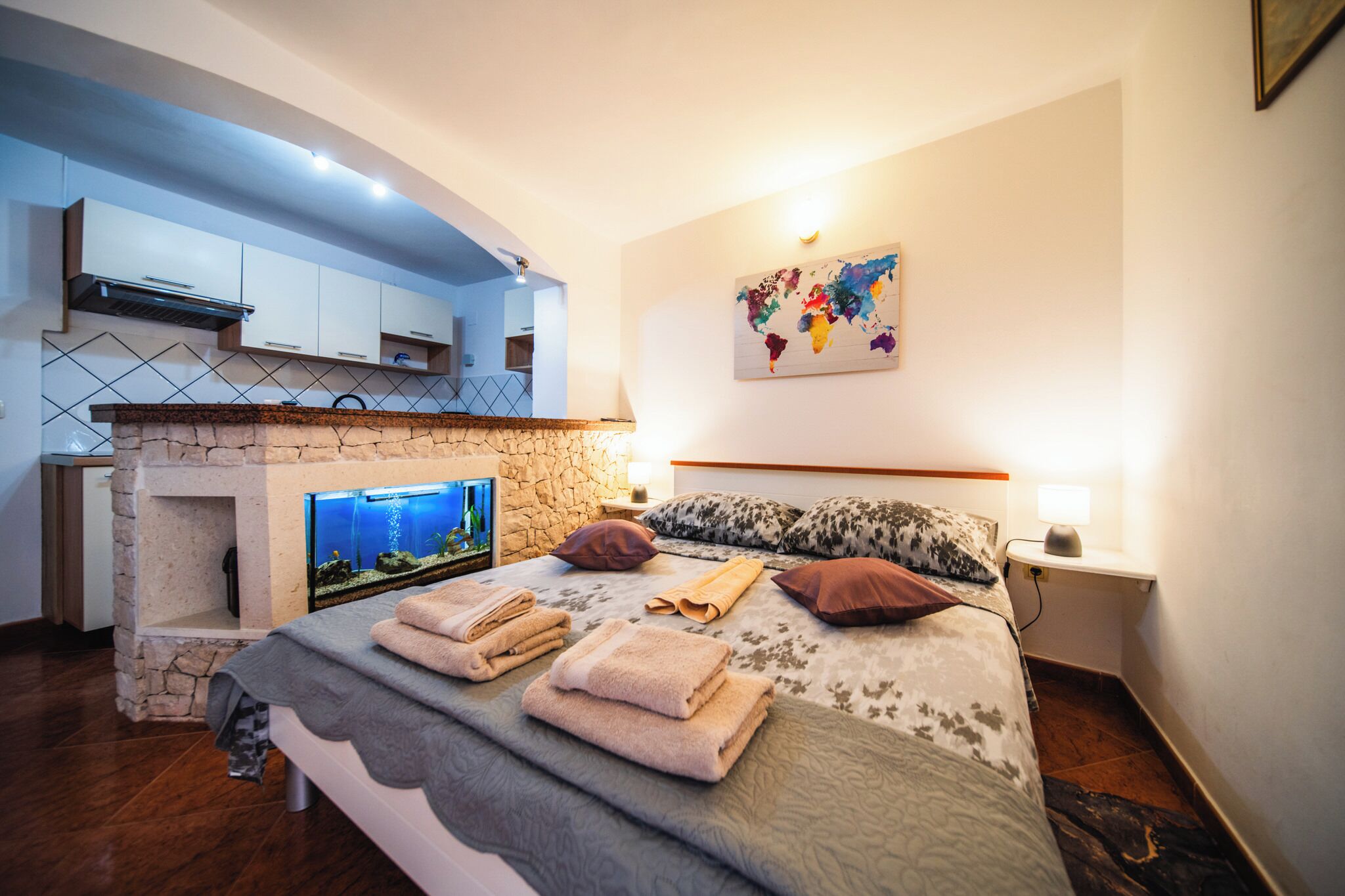 A bedroom with a stone fireplace, a bed with towels, a world map on the wall, and a kitchen area with cabinets and a stove.