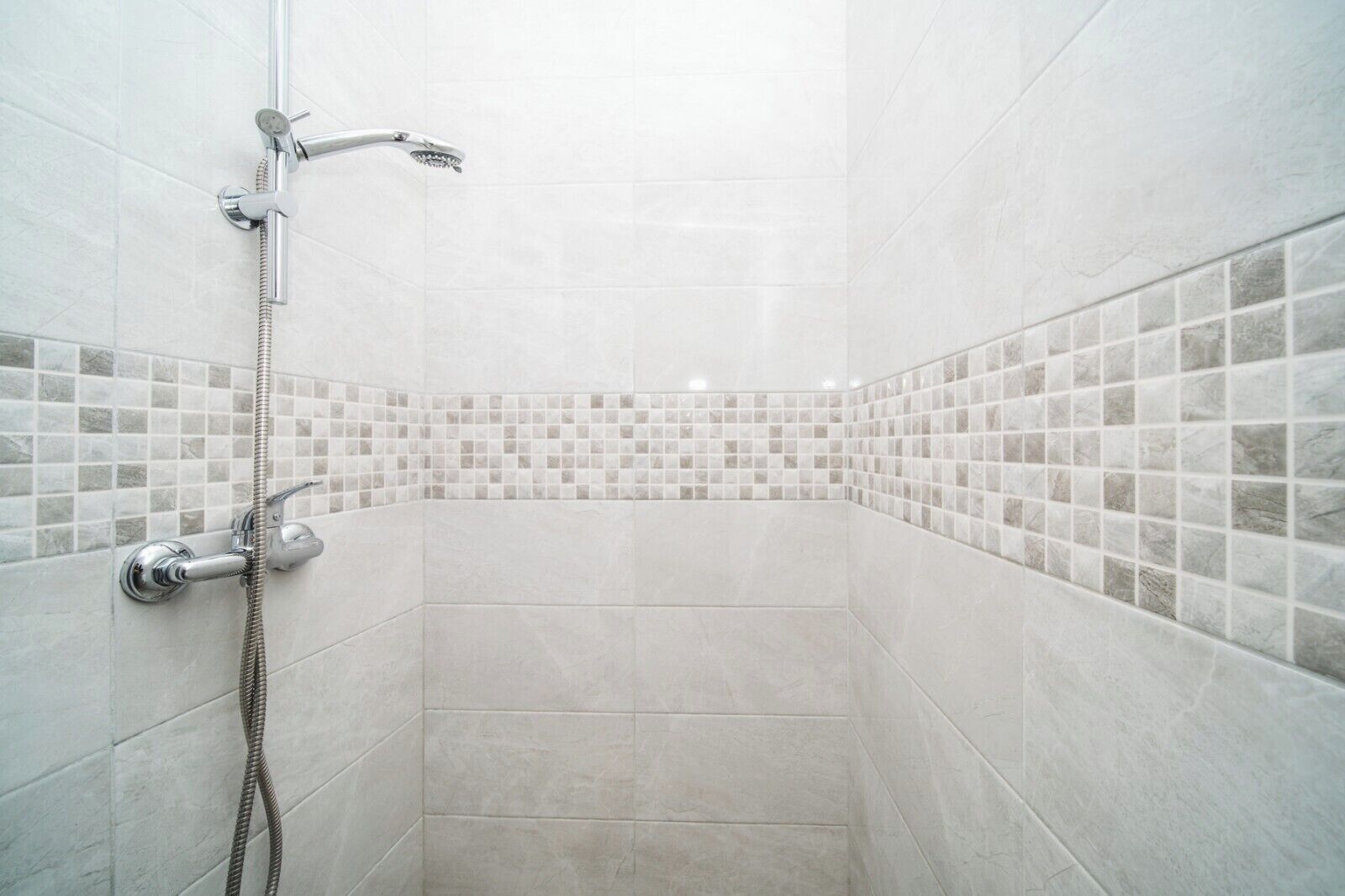 A shower with a handheld showerhead and a flexible hose, tiled walls with a mosaic border, and a corner shower design.