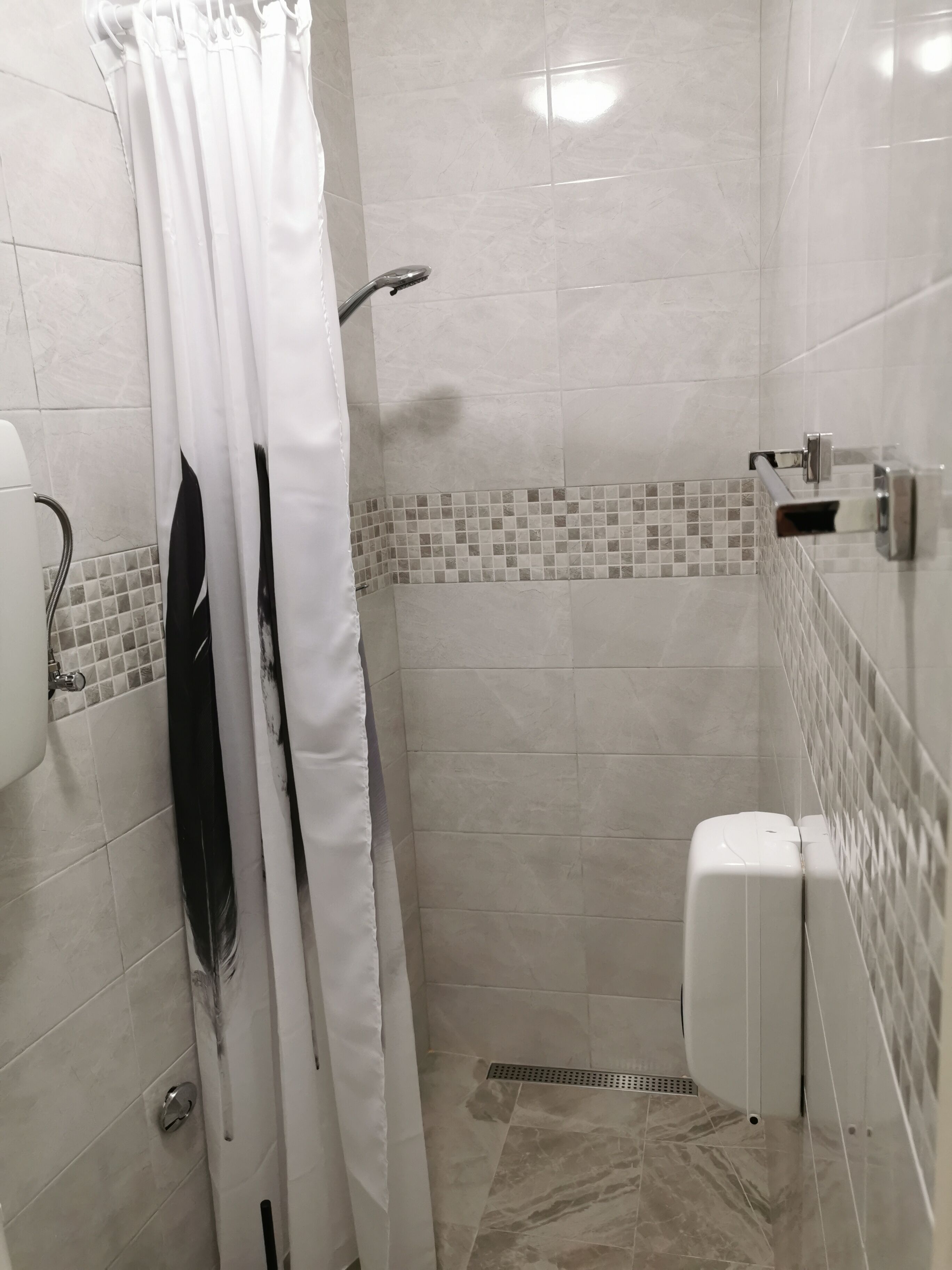 Een kleine douchecel met betegelde wanden, een witte douchegordijn, een handdouche en een wit toilet.