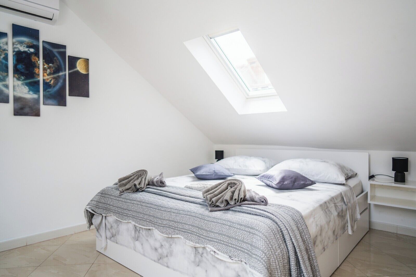 A bedroom with a sloped ceiling, a skylight, a bed with a grey and white patterned bedspread, and three framed pictures on the wall.