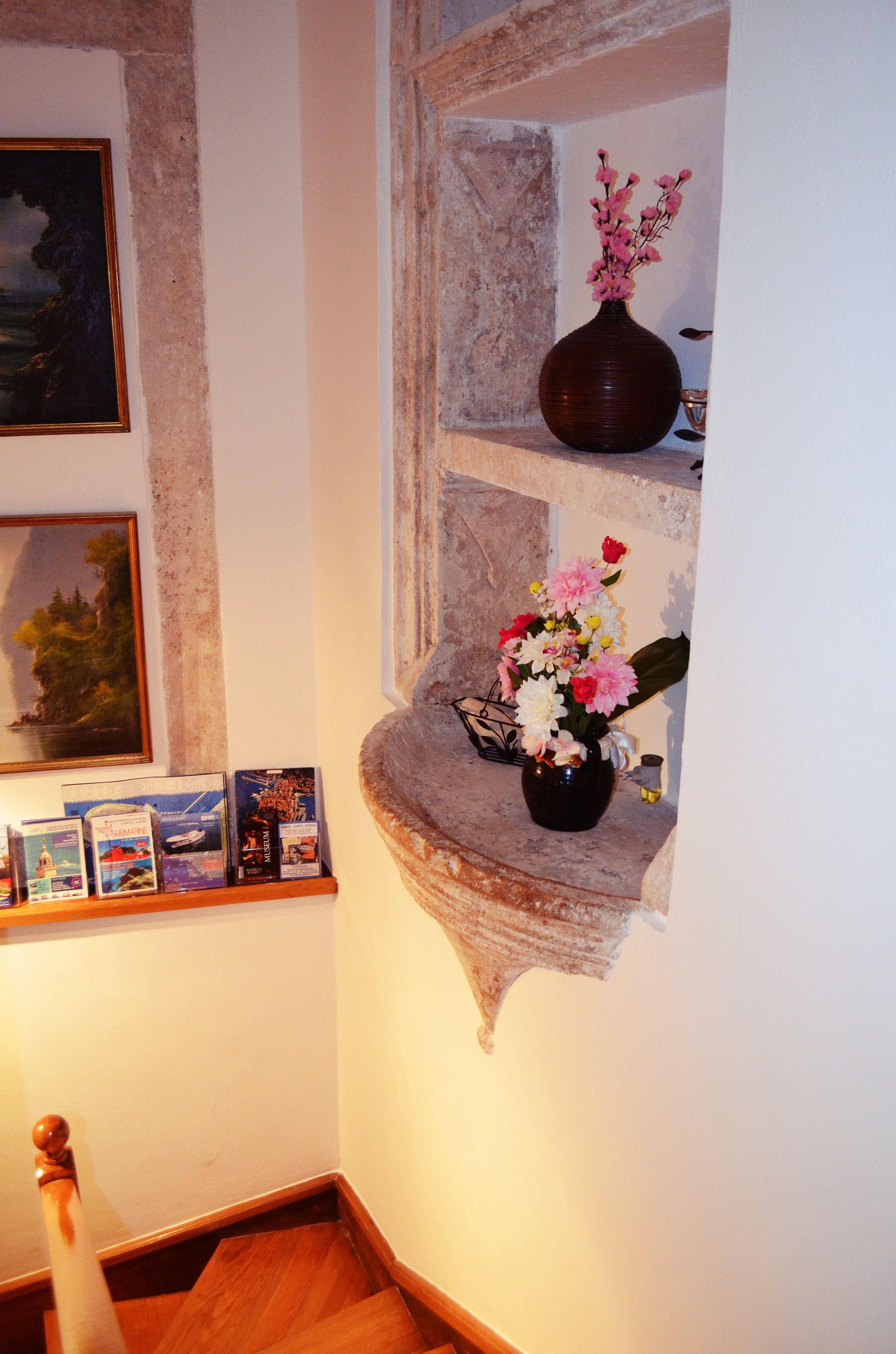 Un coin avec un escalier en bois, une étagère remplie de livres, un vase de fleurs et un tableau encadré accroché au mur.
