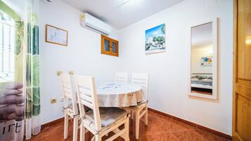 Un comedor con una mesa redonda, cuatro sillas, un aire acondicionado montado en la pared, un plano arquitectónico enmarcado y una pintura de una escena costera.