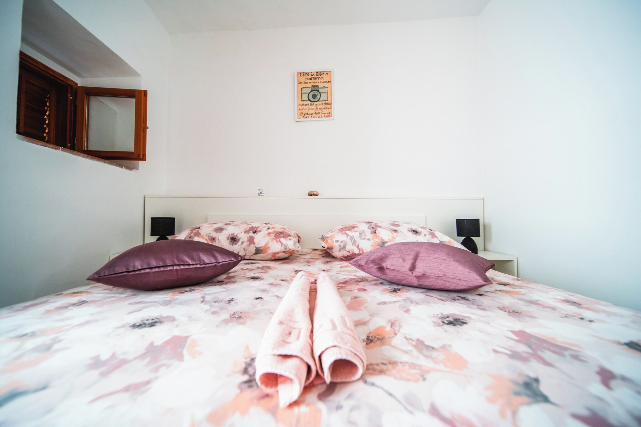 A neatly made bed with floral bedding, two pillows, and a rolled towel in a room with a white wall, a wooden window frame, and a small sign above the bed.