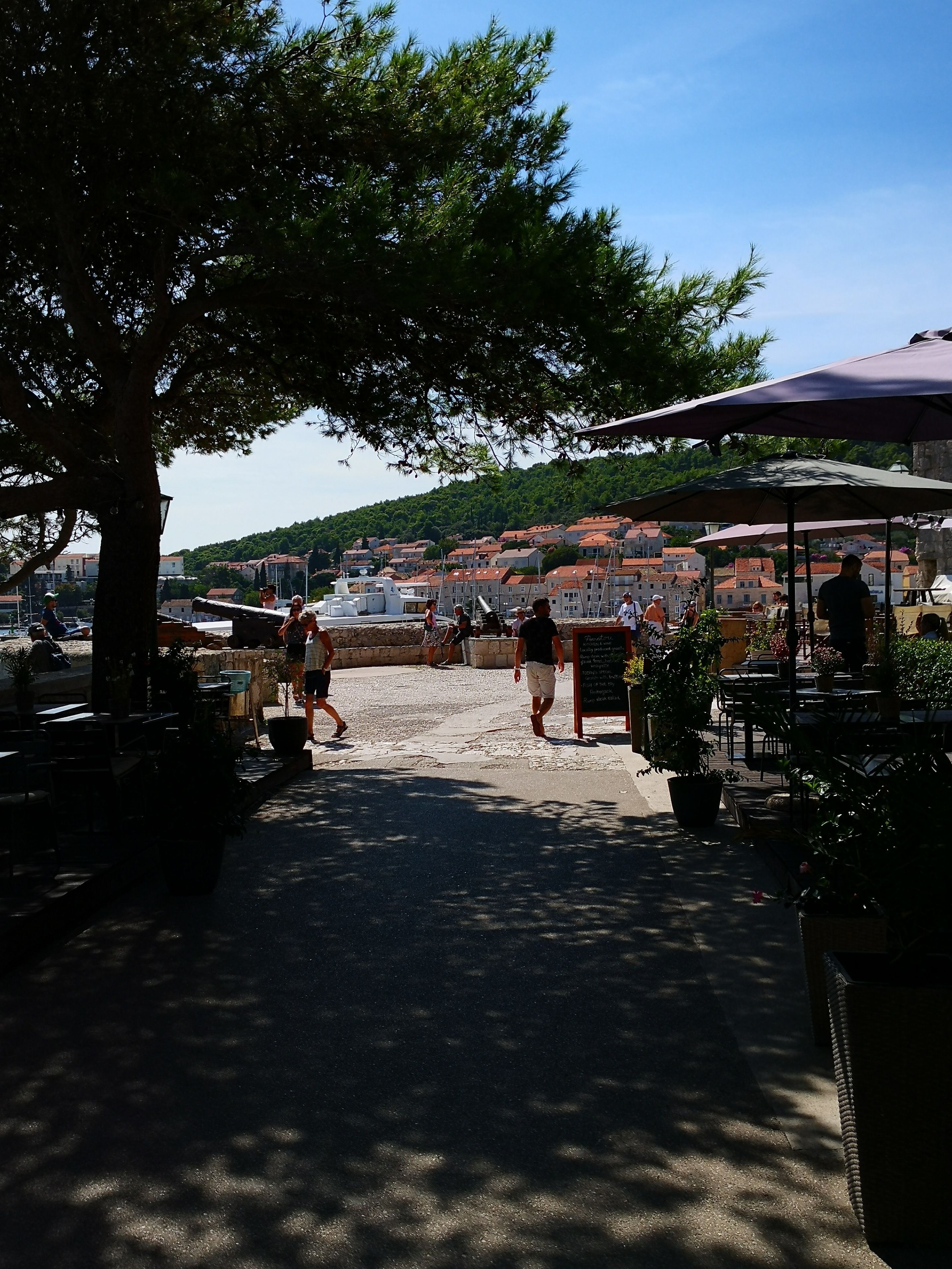 A cobblestone street with outdoor seating and umbrellas, leading to a waterfront area with buildings and greenery in the background.