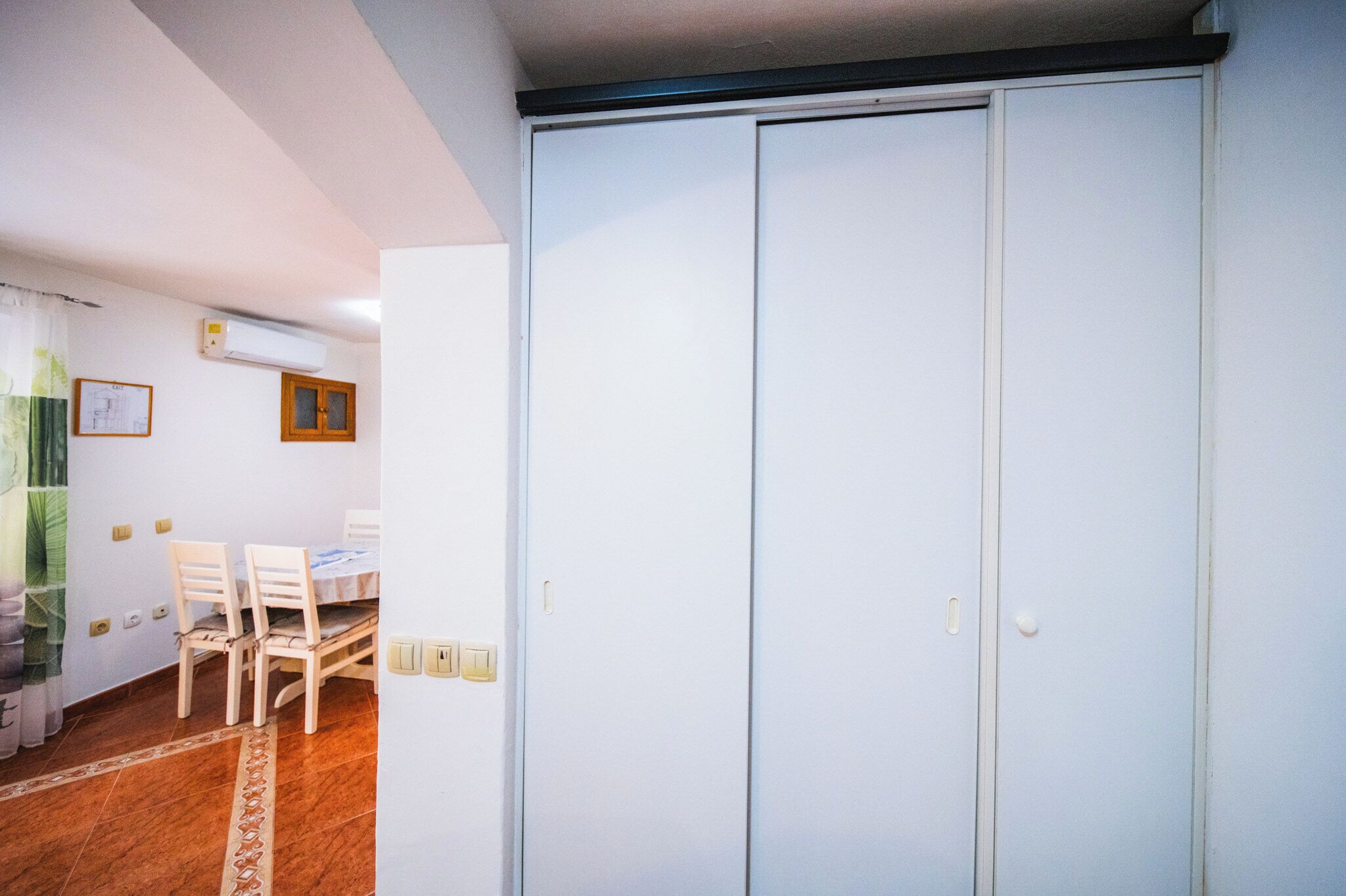 A room with a white wardrobe, a dining table with chairs, and a window with curtains.