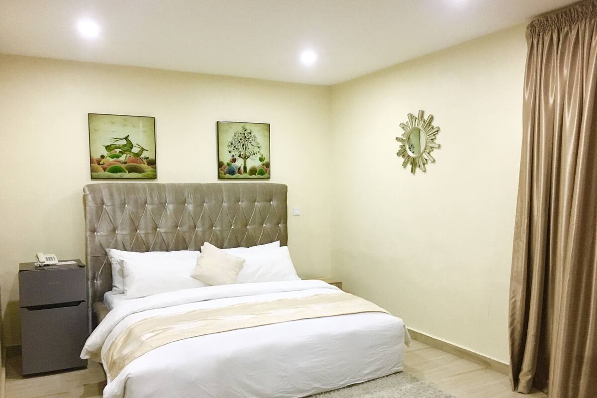 A neatly arranged bedroom with a tufted headboard, a bedside table with a phone, and two framed artworks on the wall.
