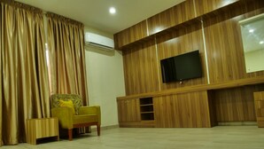 A living room with a wooden wall, a flat-screen TV mounted on the wall, a yellow armchair, and a wooden cabinet. - Alexia's Place Hotel (Lagos)