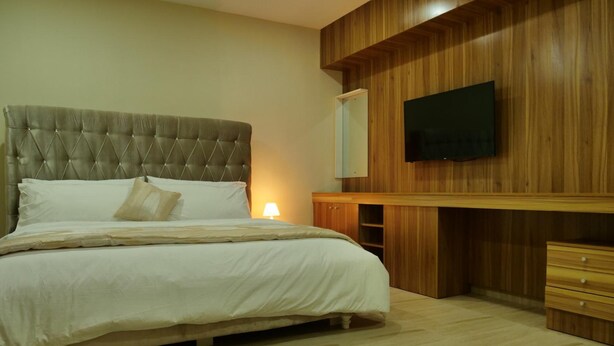 A bedroom with a tufted headboard bed, a flat-screen TV mounted on a wooden wall, and a bedside cabinet. - Alexia's Place Hotel (Lagos)