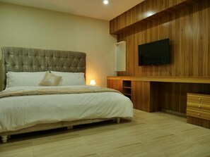 A bedroom with a tufted headboard bed, a flat-screen TV mounted on a wooden wall, and a bedside cabinet. - Alexia's Place Hotel (Lagos)