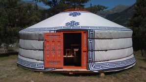 A traditional yurt with a red door and decorative patterns, set in a grassy area with trees and mountains in the background. - Yourtes de Montagnes Insolites (Val-Cenis)