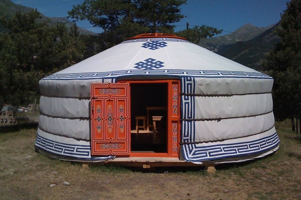 Une yourte traditionnelle avec une porte rouge et des motifs décoratifs, située dans une zone herbeuse, avec des arbres et des montagnes en arrière-plan.