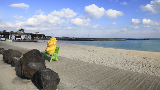 A beach with a wooden boardwalk, a green bench, and large rocks. - Daisy (Jeju City)