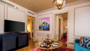 Habitación de hotel con televisor de pantalla plana, una mesa de comedor, alfombra roja estampada, un cuadro de un paisaje urbano y un sofá azul.