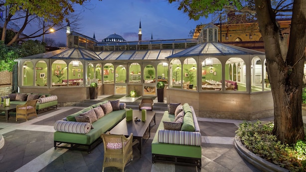 An outdoor lounge area with green seating, a glass-roofed structure, and a view of a mosque in the background.